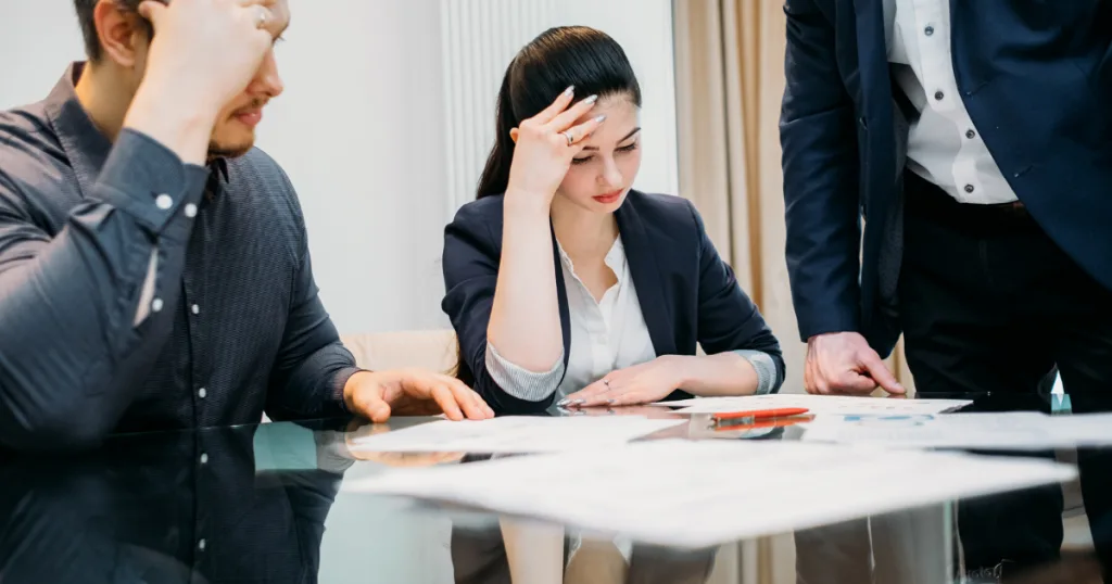 Stressed couple at a conference table reviewing papers with a standing advisor